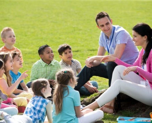 Image of woman and man talking with a class of smiling students in a park