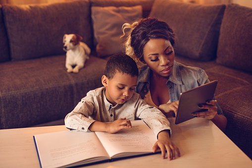 Young African American boy student of Visions In Education's Home School Academy is reading a book while his mother is using touchpad