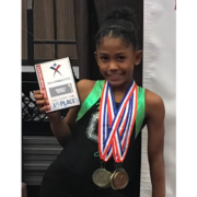Female gymnast smiling at the camera holding 1st place award with gold medals