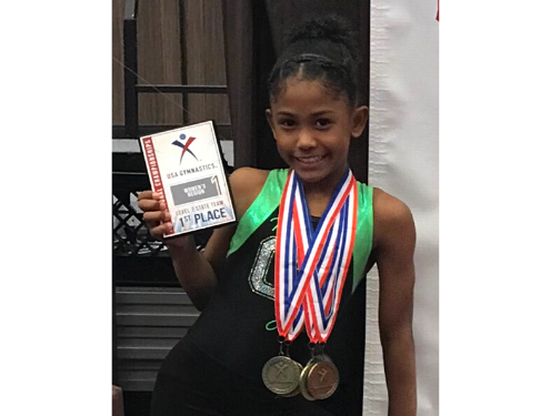 Female gymnast smiling at the camera holding 1st place award with gold medals
