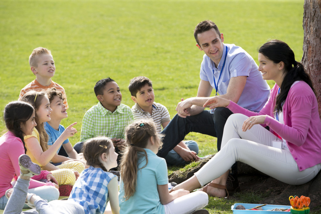 Small group of children sitting on the grass having a lesson outdoors. Male and female teacher can be seen. The children look to be listening and enjoying themselves.