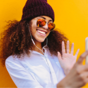 Female high school students waves into iphone taking a selfie wearing beanie and sunglasses against yellow background