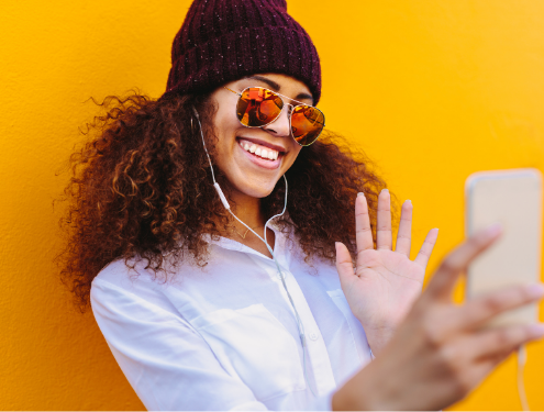 Female high school students waves into iphone taking a selfie wearing beanie and sunglasses against yellow background
