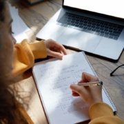 Teen girl online high school student wear headphones studying from home writing in workbook solving equations learning math sits at desk. Teenage school pupil learn online on laptop, close up over shoulder view.