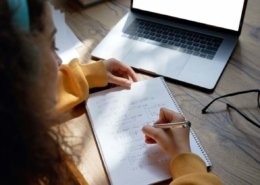 Teen girl online high school student wear headphones studying from home writing in workbook solving equations learning math sits at desk. Teenage school pupil learn online on laptop, close up over shoulder view.