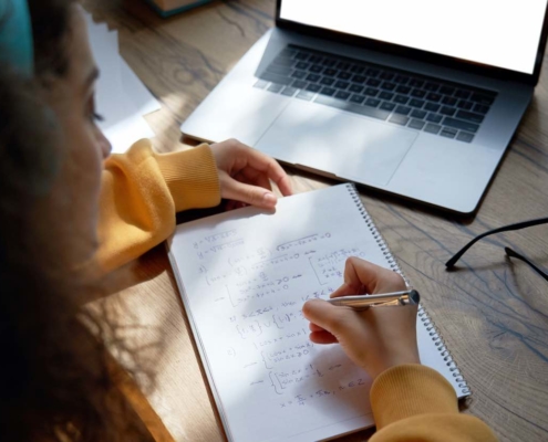 Teen girl online high school student wear headphones studying from home writing in workbook solving equations learning math sits at desk. Teenage school pupil learn online on laptop, close up over shoulder view.