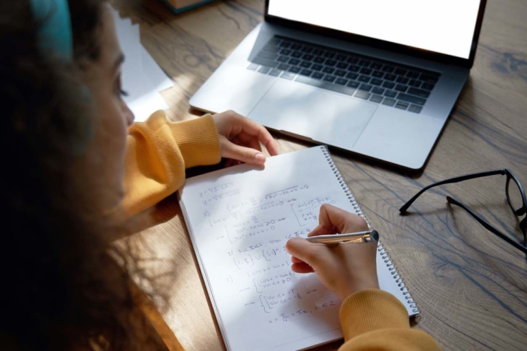 Teen girl online high school student wear headphones studying from home writing in workbook solving equations learning math sits at desk. Teenage school pupil learn online on laptop, close up over shoulder view.