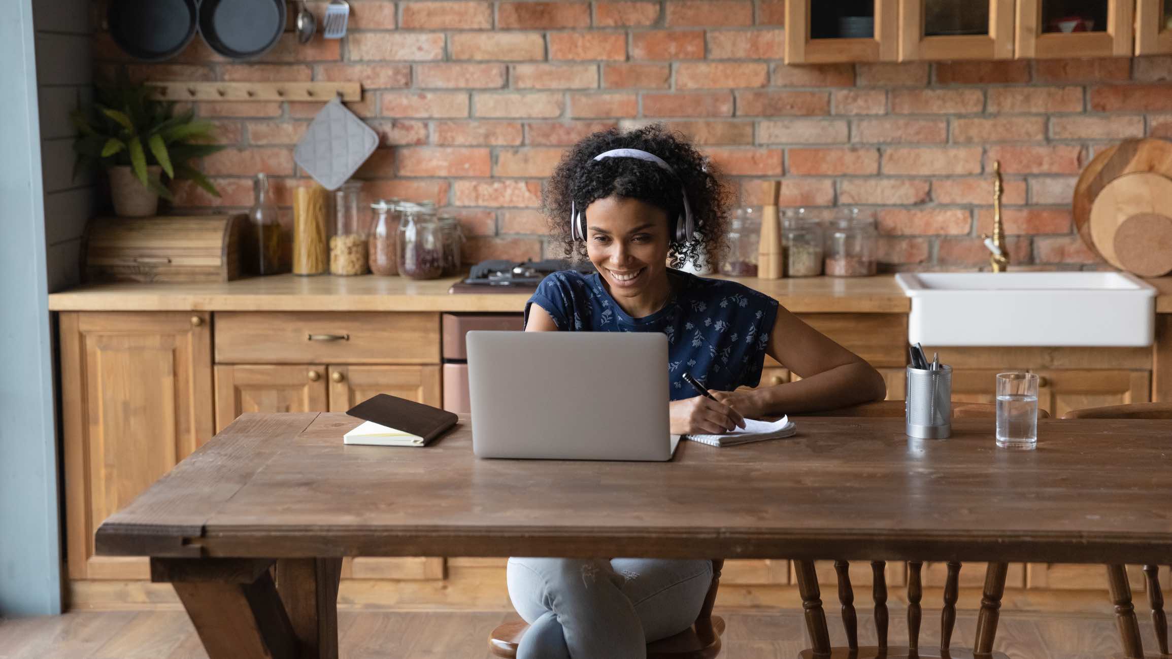 Millennial afro american woman in headphones sit by laptop engaged in distant learning foreign language at modern designed home kitchen. Smiling young black lady listen to webinar taking notes by hand