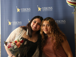 One eighth grade home school student holding a bouquet of flowers embraces her sister in front of a blue and yellow Visions In Education backdrop