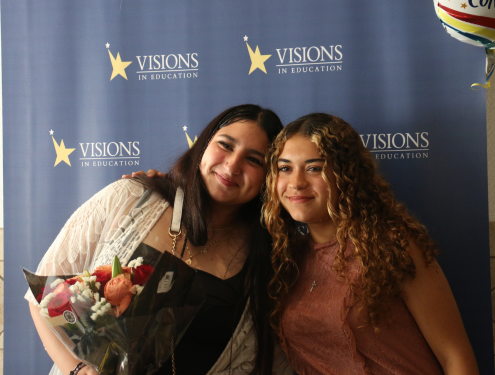 One eighth grade home school student holding a bouquet of flowers embraces her sister in front of a blue and yellow Visions In Education backdrop