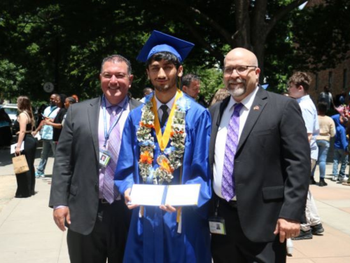 Male superintendent and principal standing on the side of male graduate in blue cap and gown outside of graduation