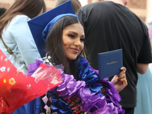 Dark haired female graduate poses with her diploma in flower leis and a cap and gown at graduation