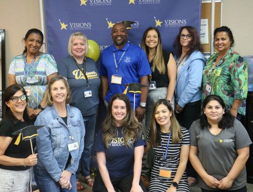 A group of male and female Visions employees smile in front of a blue, yellow and white backdrop that says Visions In Education.