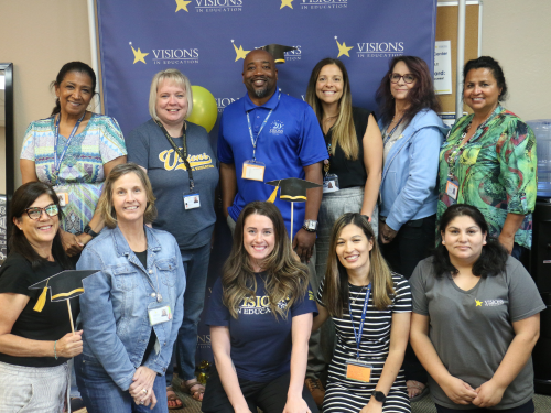 A group of male and female Visions employees smile in front of a blue, yellow and white backdrop that says Visions In Education.
