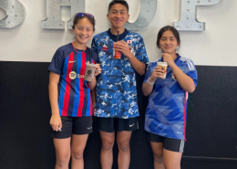 Three smiling home school students in soccer jerseys pose with Boba drinks in their hands in front of a wall at their family-owned cookie business
