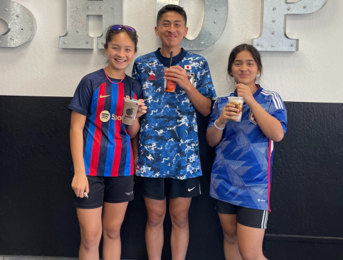 Three smiling home school students in soccer jerseys pose with Boba drinks in their hands in front of a wall at their family-owned cookie business