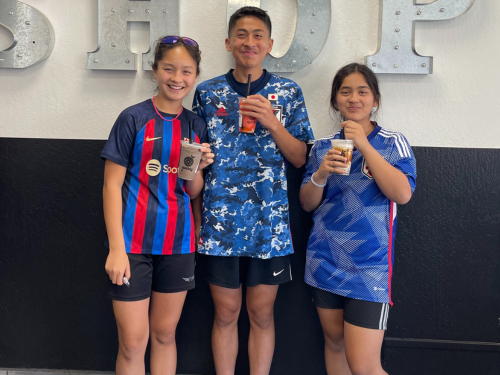 Three smiling home school students in soccer jerseys pose with Boba drinks in their hands in front of a wall at their family-owned cookie business