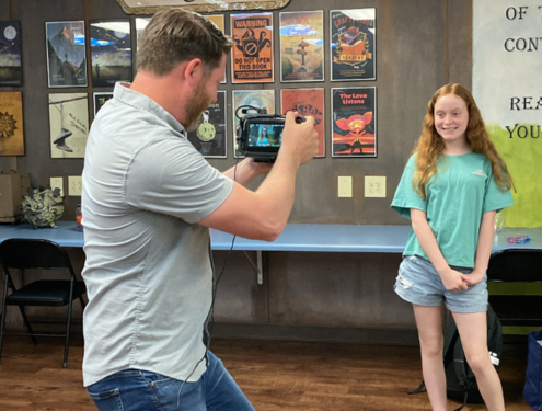 Camera man records smiling Visions student standing in front of a wall of published writing