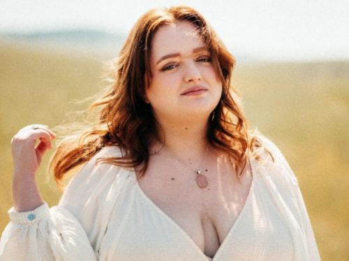 Redheaded high school female in a white dress poses in an open field for graduation pictures