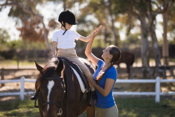 Side view of woman giving high five to girl sitting on horse in paddock, taking horseback riding lessons for enrichment activity