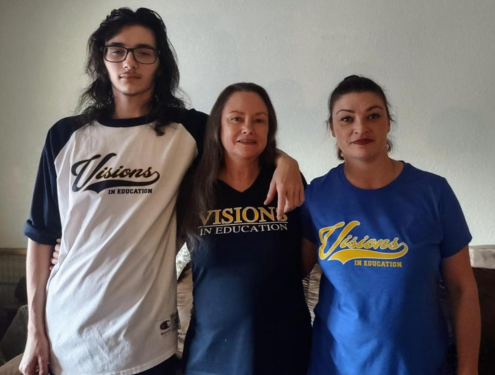 A teenage boy, his grandmother and his mother stand in a row in different Visions In Education shirts and smile for the camera.