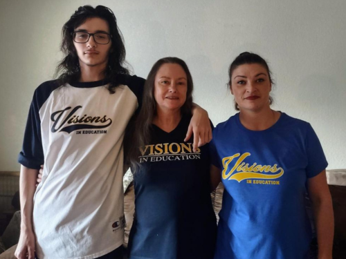 A teenage boy, his grandmother and his mother stand in a row in different Visions In Education shirts and smile for the camera.