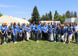 Large group of Visions staff in matching light blue t-shirts stand together on a lawn and smile for a photo.