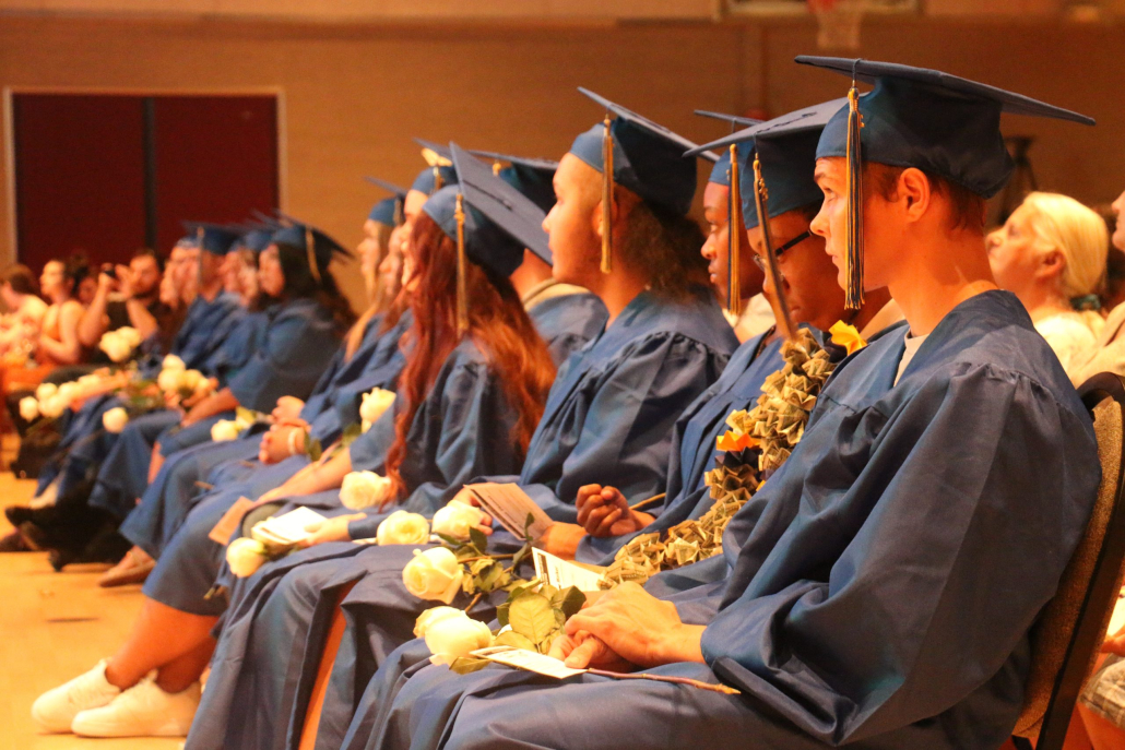 A line of sitting graduates in blue caps and gowns holding white roses.