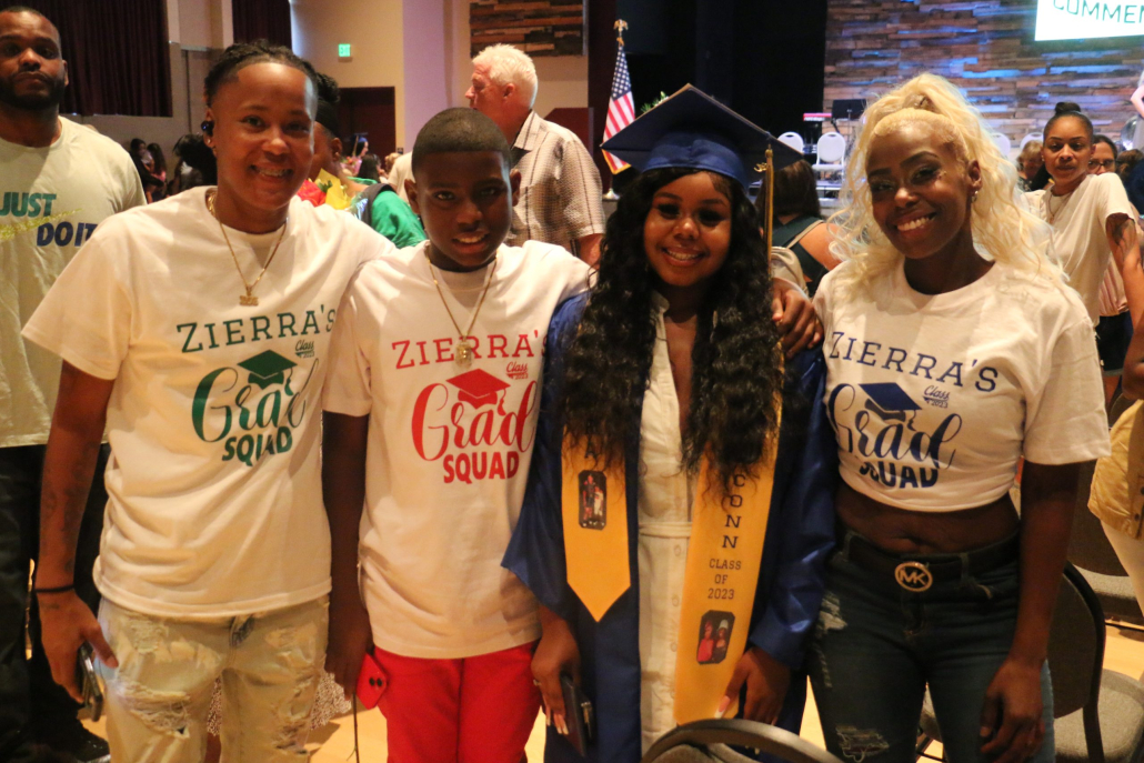 A graduate in a blue cap and gown stands with her three family members in matching "Zierra's Grad Squad" t-shirts.