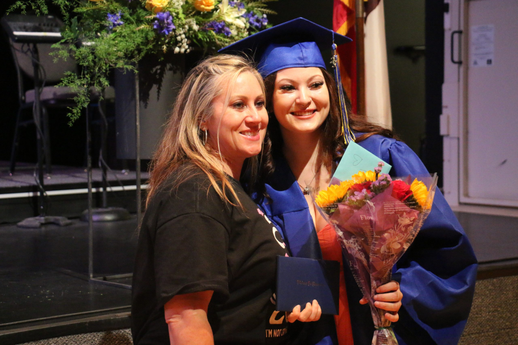 A graduate in a blue cap and gown and a bouquet of flowers in her hand stands smiling next to her mom.