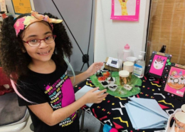 Nine year old girl with glasses and a floral headband smiles and points to her colorful artwork displayed on a table