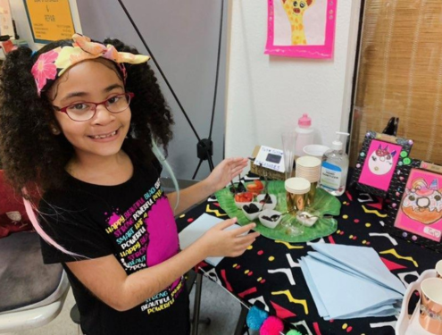 Nine year old girl with glasses and a floral headband smiles and points to her colorful artwork displayed on a table