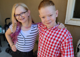 Two young siblings, a girl with glasses and a black and white striped shirt and a boy in a red and white checkered shirt, stand next to one another and smile for a first day of school photo.