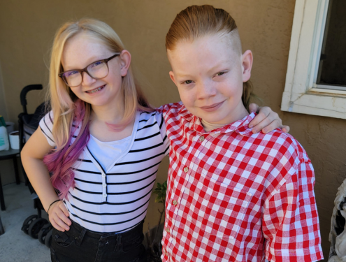 Two young siblings, a girl with glasses and a black and white striped shirt and a boy in a red and white checkered shirt, stand next to one another and smile for a first day of school photo.
