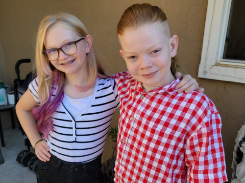 Two young siblings, a girl with glasses and a black and white striped shirt and a boy in a red and white checkered shirt, stand next to one another and smile for a first day of school photo.