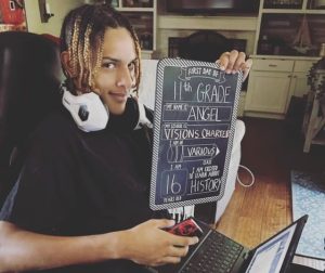 Black teen poses at home on the first day of independent study high school with a sign, laptop and headphones