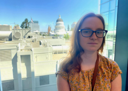 Smiling girl with glasses in a yellow shirt stands in front of a window overlooking downtown Sacramento, California