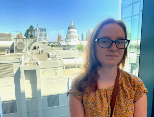 Smiling girl with glasses in a yellow shirt stands in front of a window overlooking downtown Sacramento, California