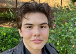 Selfie of a teenage boy with brown hair wearing a leather jacket in front of green bushes.