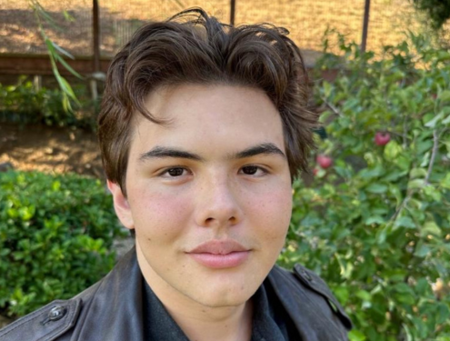 Selfie of a teenage boy with brown hair wearing a leather jacket in front of green bushes.