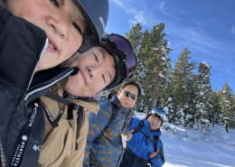 A family of four smile for a photo in snow gear as they ride on the chair lift of a ski resort.