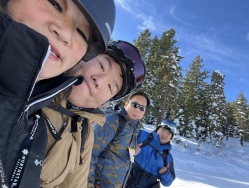 A family of four smile for a photo in snow gear as they ride on the chair lift of a ski resort.