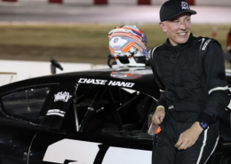 Chase Hand, wearing a black had and all-black racing suit, leans against a black race car with his name and number 27 on it.