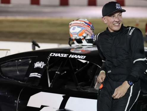 Chase Hand, wearing a black had and all-black racing suit, leans against a black race car with his name and number 27 on it.