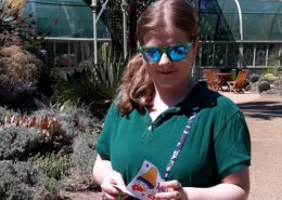 Teenage girl with a side ponytail wearing blue reflective sunglasses and a purse diagonally across her body looks down at a pamphlet in her hands.