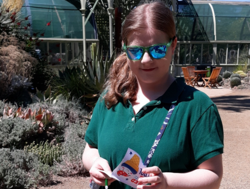 Teenage girl with a side ponytail wearing blue reflective sunglasses and a purse diagonally across her body looks down at a pamphlet in her hands.