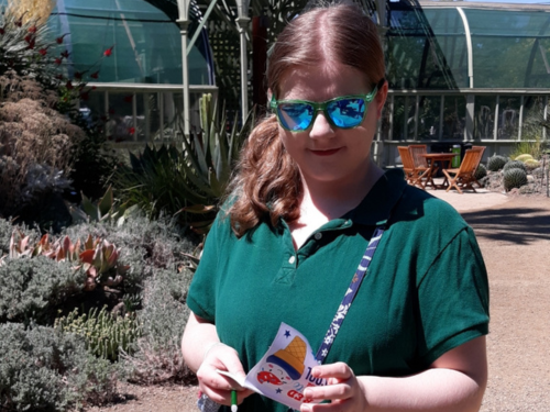 Teenage girl with a side ponytail wearing blue reflective sunglasses and a purse diagonally across her body looks down at a pamphlet in her hands.