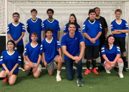 Visions students and soccer club members in blue jerseys gather in front of the soccer net on the turf for a group photo.