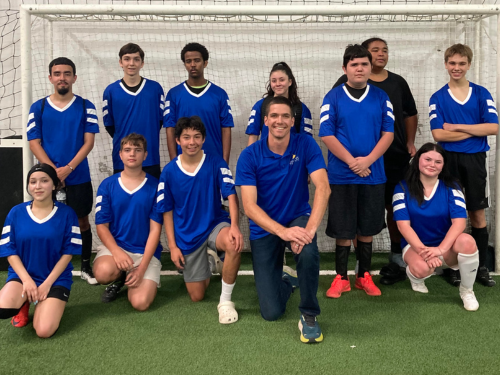 Visions students and soccer club members in blue jerseys gather in front of the soccer net on the turf for a group photo.