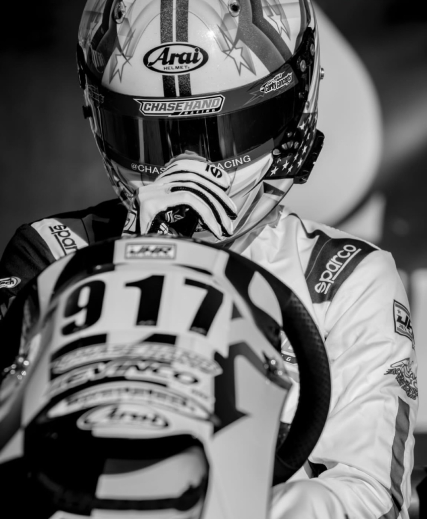 A black and white photo of Chase wearing his racing gear sitting on a go kart with the number 917 on it getting ready to begin his race.